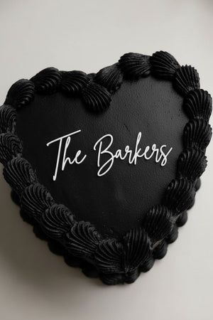 Wedding cake toppers personalized acrylic last name sign “The Barkers” on black heart shaped wedding cake with textured frosting on white background