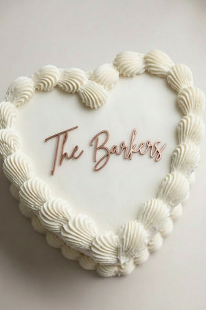Wedding cake toppers personalized acrylic last name sign “The Barkers” on white heart shaped wedding cake with piped frosting top view on light background