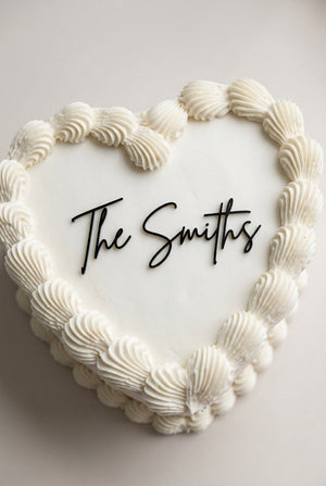 Wedding cake toppers personalized acrylic name sign “The Smiths” on heart shaped wedding cake with piped frosting on light background