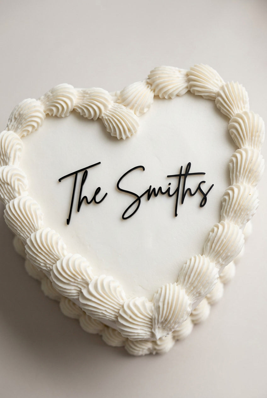 Wedding cake toppers personalized acrylic name sign “The Smiths” on heart shaped wedding cake with piped frosting on light background